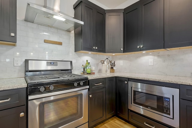 kitchen with decorative backsplash, wall chimney exhaust hood, light stone counters, stainless steel appliances, and light hardwood / wood-style flooring