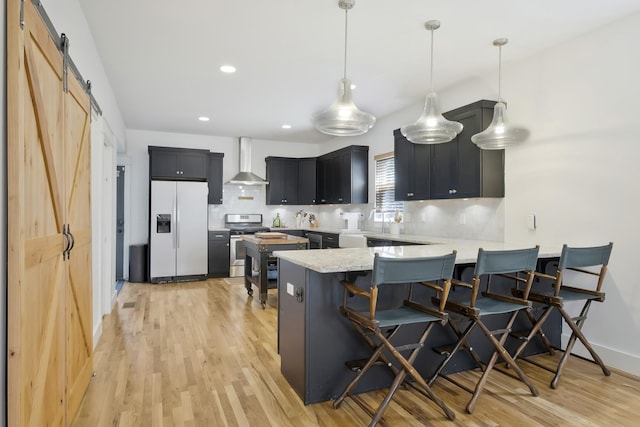 kitchen with stainless steel stove, decorative light fixtures, white refrigerator with ice dispenser, a barn door, and wall chimney range hood