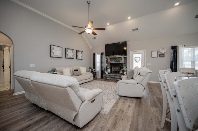 living room with ceiling fan, hardwood / wood-style floors, high vaulted ceiling, ornamental molding, and a stone fireplace