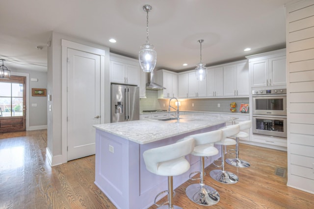 kitchen featuring pendant lighting, white cabinetry, stainless steel appliances, light stone counters, and an island with sink