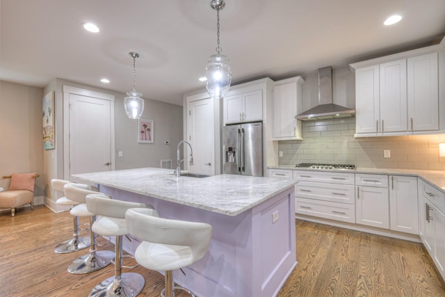 kitchen with wall chimney range hood, sink, stainless steel appliances, white cabinets, and a center island with sink