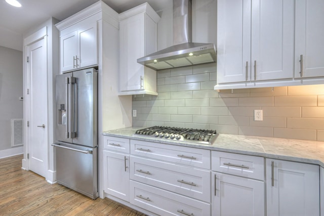 kitchen featuring appliances with stainless steel finishes, white cabinetry, light hardwood / wood-style floors, light stone counters, and wall chimney exhaust hood