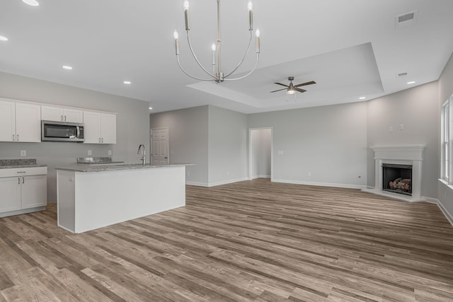 kitchen with white cabinets, a raised ceiling, light stone countertops, a center island with sink, and light hardwood / wood-style flooring