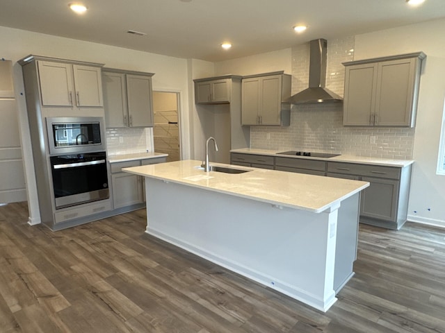 kitchen featuring sink, gray cabinets, an island with sink, stainless steel appliances, and wall chimney range hood