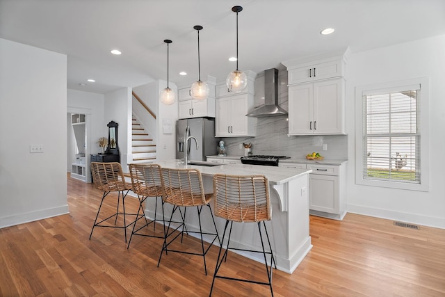 kitchen with stainless steel fridge, white cabinets, a kitchen bar, a kitchen island with sink, and wall chimney range hood