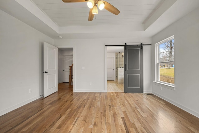 unfurnished bedroom with ceiling fan, a barn door, light hardwood / wood-style floors, and a tray ceiling