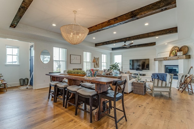 dining area featuring beamed ceiling, a large fireplace, ceiling fan with notable chandelier, and light hardwood / wood-style flooring