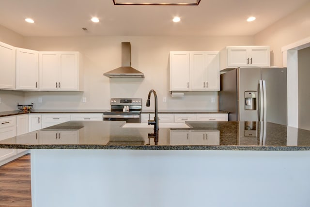 kitchen with white cabinets, dark stone counters, a kitchen island with sink, stainless steel appliances, and wall chimney range hood