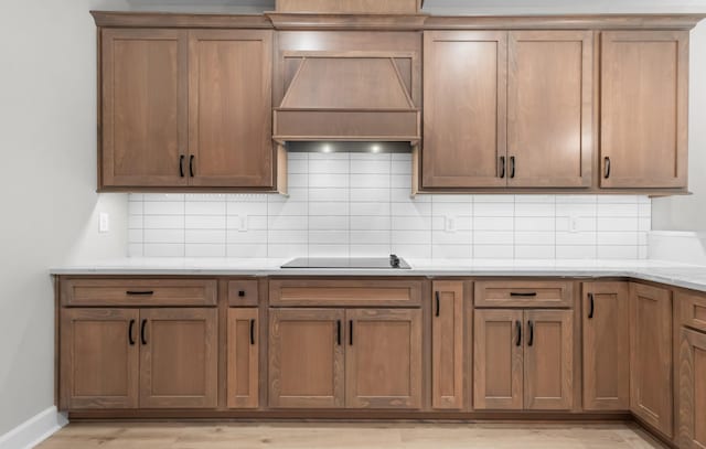 kitchen featuring backsplash, custom exhaust hood, black electric stovetop, light stone counters, and light hardwood / wood-style floors