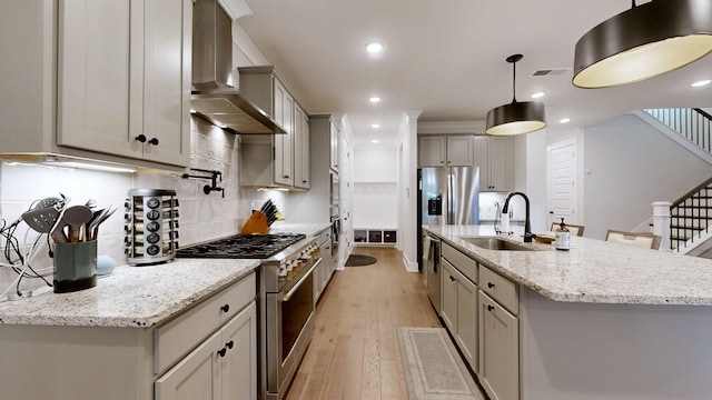 kitchen featuring appliances with stainless steel finishes, sink, a kitchen island with sink, light stone counters, and wall chimney exhaust hood