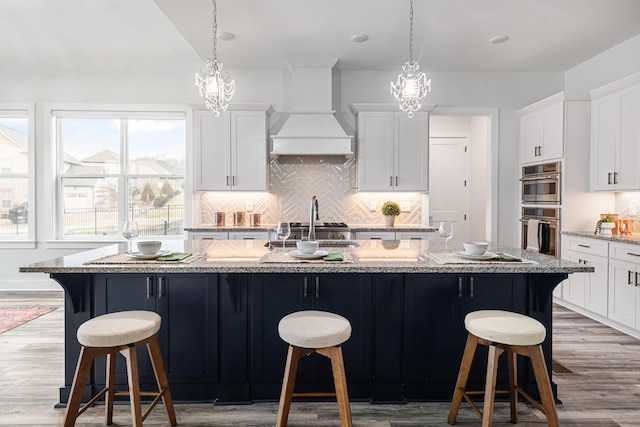 kitchen with custom range hood, hanging light fixtures, an island with sink, and stainless steel double oven