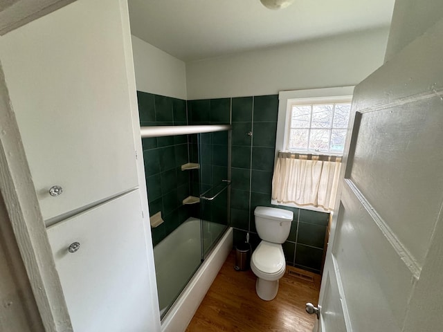 bathroom with bath / shower combo with glass door, wood finished floors, toilet, and tile walls