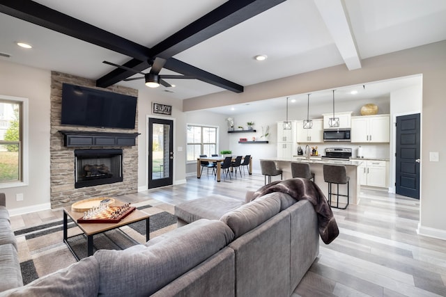 living room with beam ceiling, ceiling fan, a stone fireplace, and light hardwood / wood-style floors