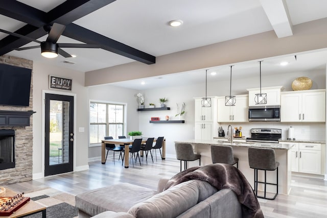 living room featuring a stone fireplace, sink, ceiling fan, beam ceiling, and light hardwood / wood-style floors