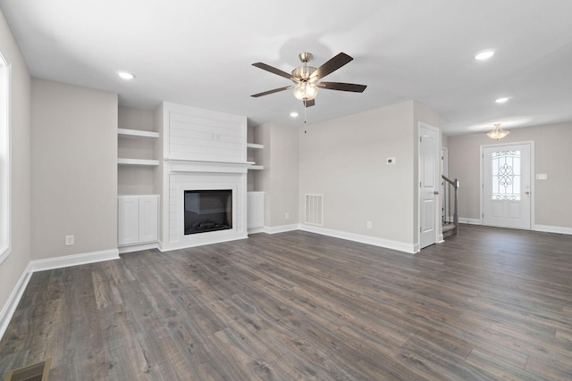 unfurnished living room with dark wood-type flooring, a large fireplace, ceiling fan, and built in shelves