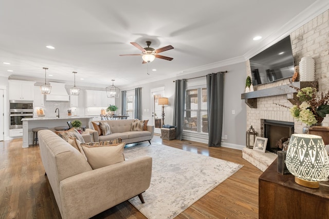 living room with sink, ornamental molding, light hardwood / wood-style floors, a brick fireplace, and ceiling fan with notable chandelier