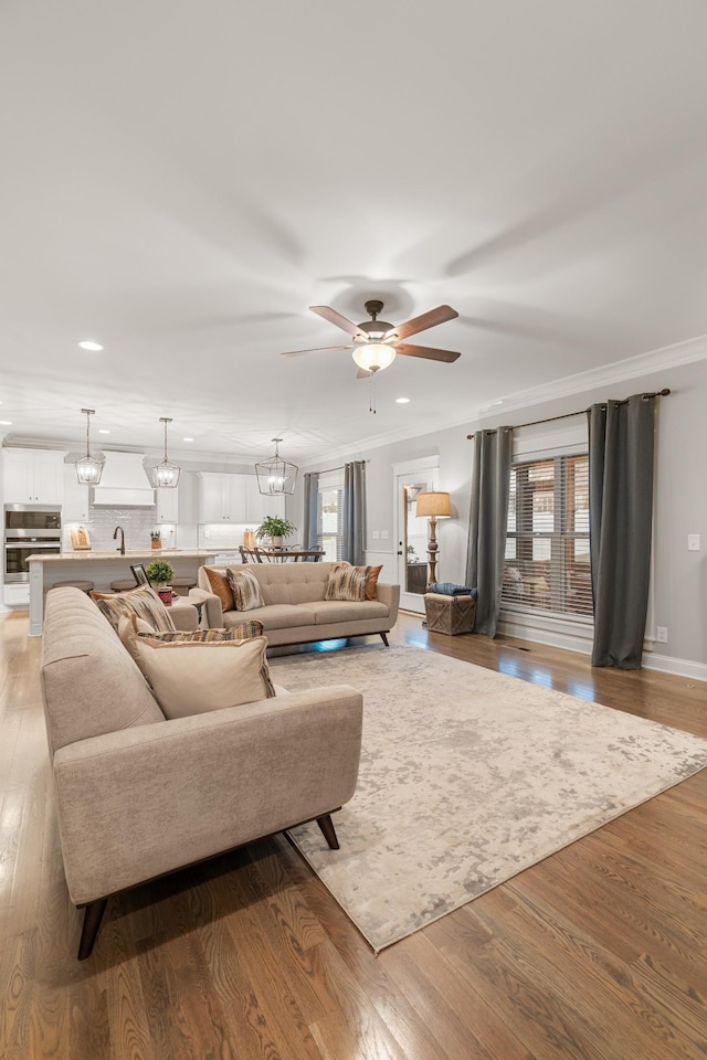 living room with ornamental molding, sink, ceiling fan with notable chandelier, and light wood-type flooring