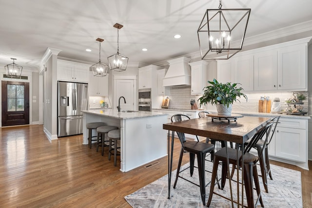 kitchen featuring pendant lighting, premium range hood, stainless steel refrigerator with ice dispenser, an island with sink, and white cabinets