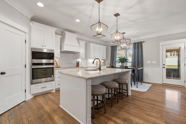 kitchen featuring sink, custom exhaust hood, a center island with sink, pendant lighting, and white cabinets