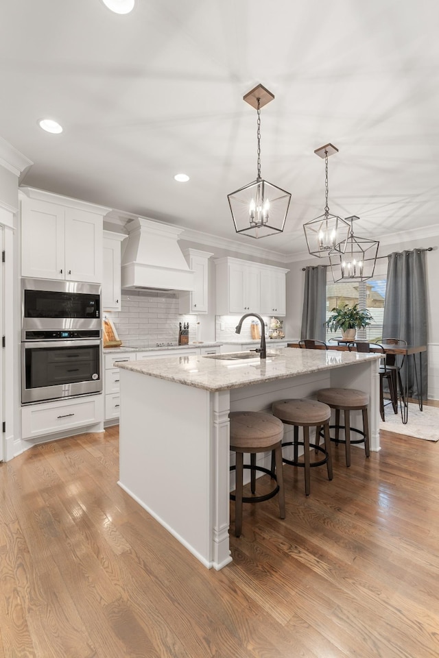 kitchen with white cabinetry, sink, custom range hood, and a center island with sink