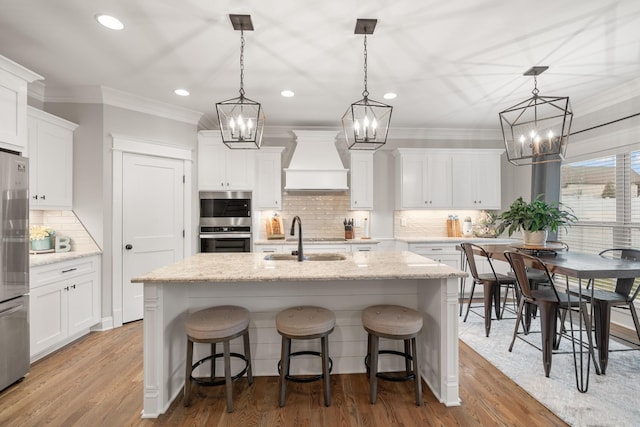 kitchen featuring white cabinetry, sink, custom exhaust hood, hanging light fixtures, and a center island with sink