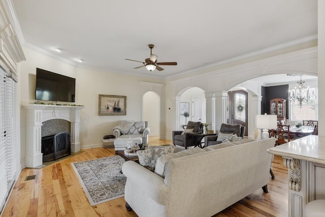 living area with arched walkways, ceiling fan with notable chandelier, a fireplace with flush hearth, light wood-style floors, and crown molding