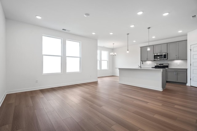 kitchen featuring gray cabinets, appliances with stainless steel finishes, decorative light fixtures, wood-type flooring, and a kitchen island with sink