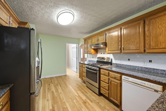 kitchen with dark stone countertops, backsplash, stainless steel appliances, a textured ceiling, and light wood-type flooring