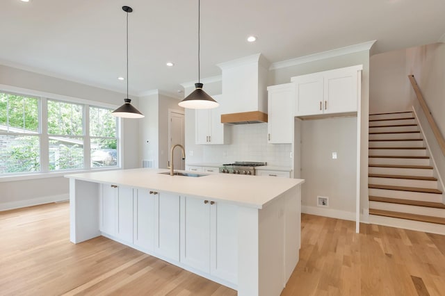 kitchen featuring sink, premium range hood, a kitchen island with sink, hanging light fixtures, and white cabinets