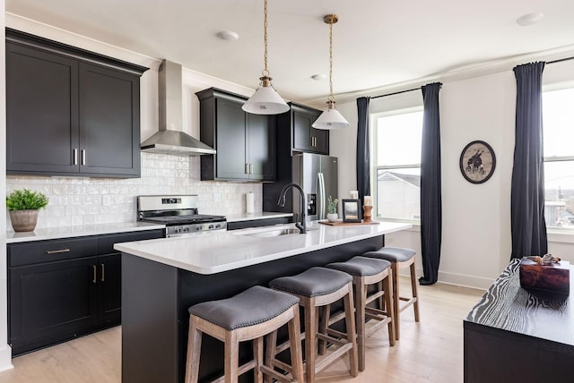 kitchen with wall chimney range hood, sink, appliances with stainless steel finishes, a center island with sink, and a kitchen bar