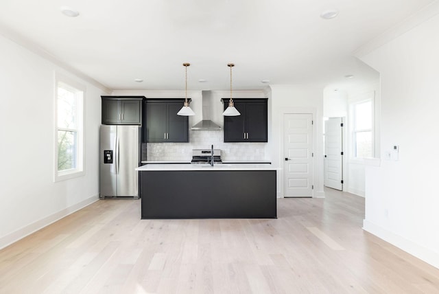 kitchen featuring hanging light fixtures, stainless steel appliances, a kitchen island with sink, decorative backsplash, and wall chimney range hood