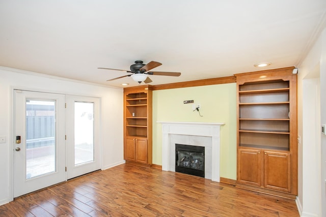 unfurnished living room with a tile fireplace, built in features, hardwood / wood-style flooring, ceiling fan, and crown molding