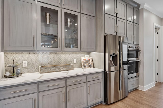 kitchen featuring light wood-type flooring, gray cabinets, stainless steel appliances, light stone countertops, and decorative backsplash