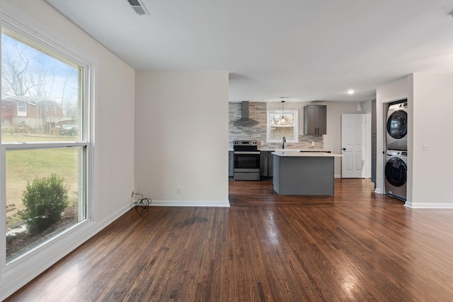 kitchen with gray cabinetry, stainless steel range with electric cooktop, light countertops, stacked washing maching and dryer, and wall chimney exhaust hood