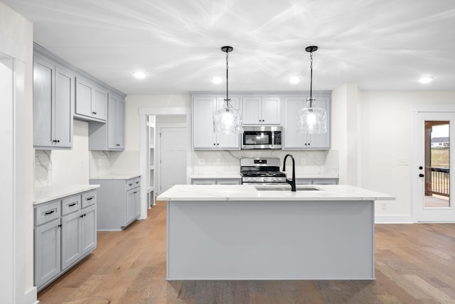 kitchen with light wood finished floors, appliances with stainless steel finishes, a kitchen island with sink, gray cabinetry, and a sink