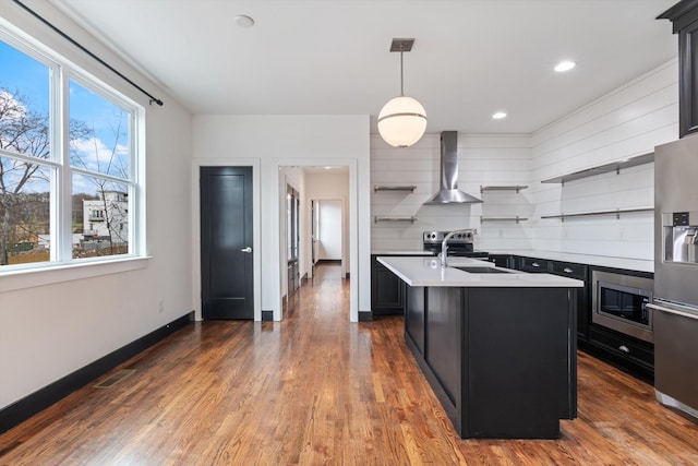 kitchen featuring sink, wall chimney range hood, hanging light fixtures, stainless steel appliances, and an island with sink