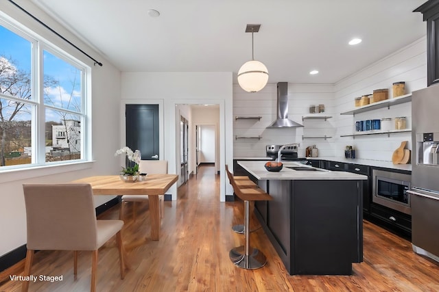 kitchen featuring decorative light fixtures, an island with sink, light hardwood / wood-style floors, stainless steel appliances, and wall chimney range hood