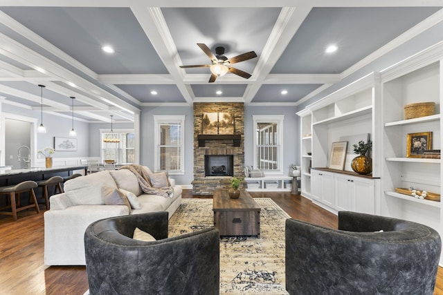 living area featuring dark wood-type flooring, beam ceiling, a stone fireplace, ceiling fan with notable chandelier, and coffered ceiling
