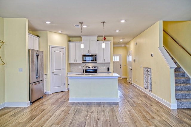 kitchen with appliances with stainless steel finishes, white cabinetry, an island with sink, hanging light fixtures, and light hardwood / wood-style flooring