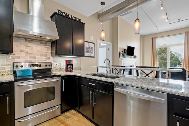 kitchen with sink, stainless steel appliances, light stone counters, decorative light fixtures, and wall chimney exhaust hood