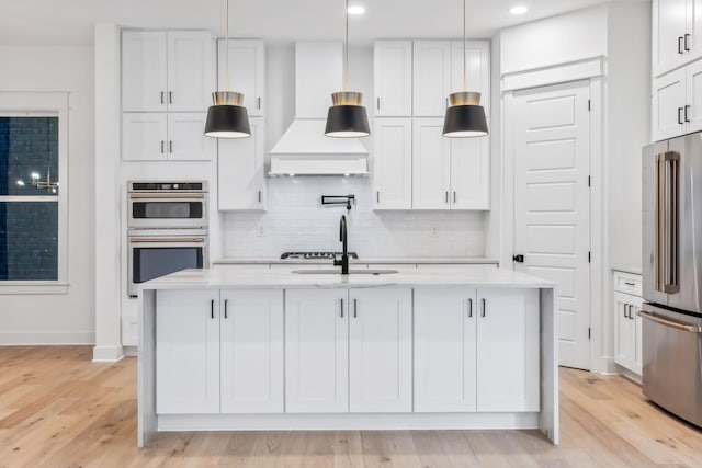 kitchen with high quality fridge, double oven, white cabinetry, an island with sink, and custom range hood