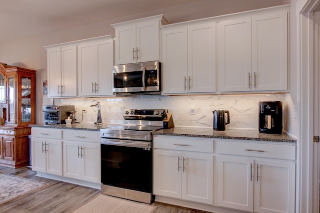 kitchen with light wood finished floors, dark stone counters, appliances with stainless steel finishes, white cabinetry, and backsplash