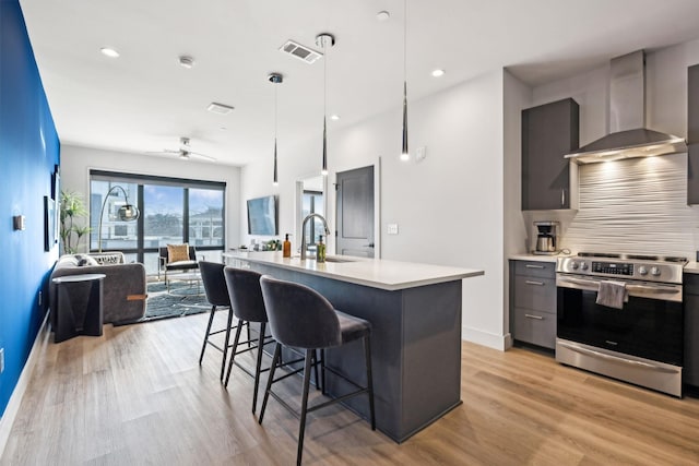 kitchen featuring pendant lighting, sink, stainless steel range with electric stovetop, a kitchen island with sink, and wall chimney range hood