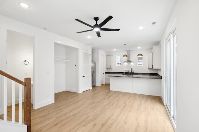 kitchen with wall chimney range hood, light hardwood / wood-style flooring, appliances with stainless steel finishes, white cabinets, and decorative light fixtures