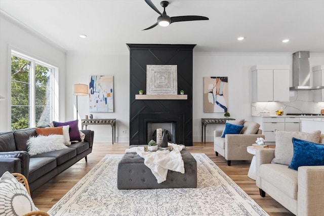 living room featuring ceiling fan, a large fireplace, crown molding, and light wood-type flooring