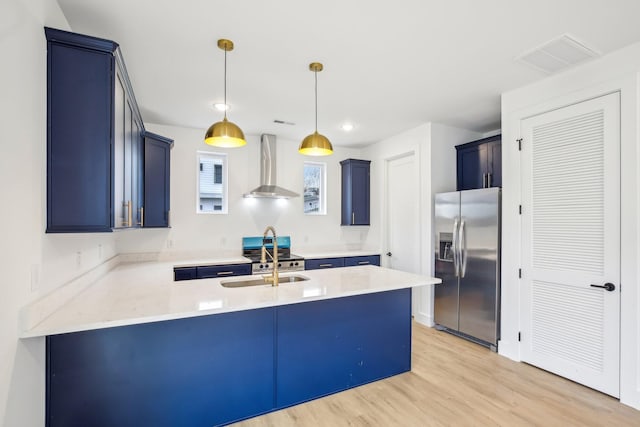 kitchen featuring appliances with stainless steel finishes, decorative light fixtures, sink, wall chimney range hood, and light wood-type flooring