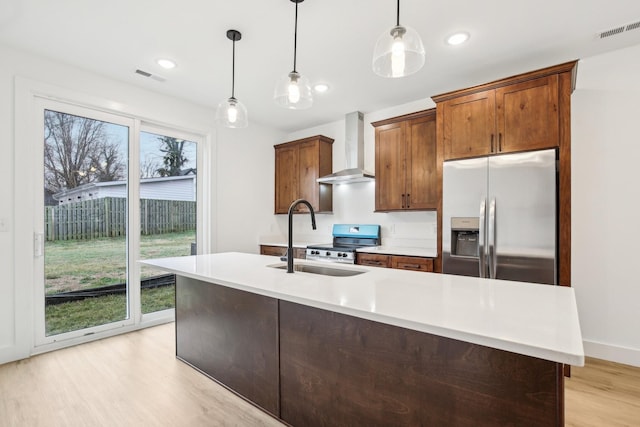 kitchen with wall chimney exhaust hood, sink, hanging light fixtures, appliances with stainless steel finishes, and an island with sink