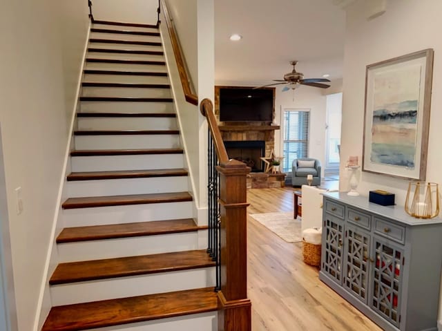 stairway with hardwood / wood-style flooring, ceiling fan, and a stone fireplace