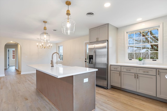 kitchen with sink, decorative light fixtures, light wood-type flooring, stainless steel fridge, and an island with sink