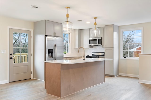 kitchen featuring appliances with stainless steel finishes, tasteful backsplash, an island with sink, gray cabinetry, and a healthy amount of sunlight